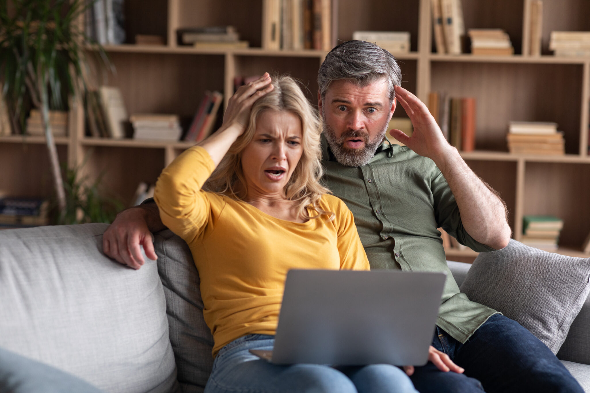 Online Scam. Emotional middle aged couple looking at laptop screen with shock, frustrated upset spouses suffering problems with computer while sitting together on couch at home, free space